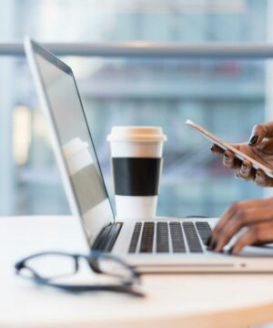 person sits at table typing on a laptop while scrolling on phone getting updates