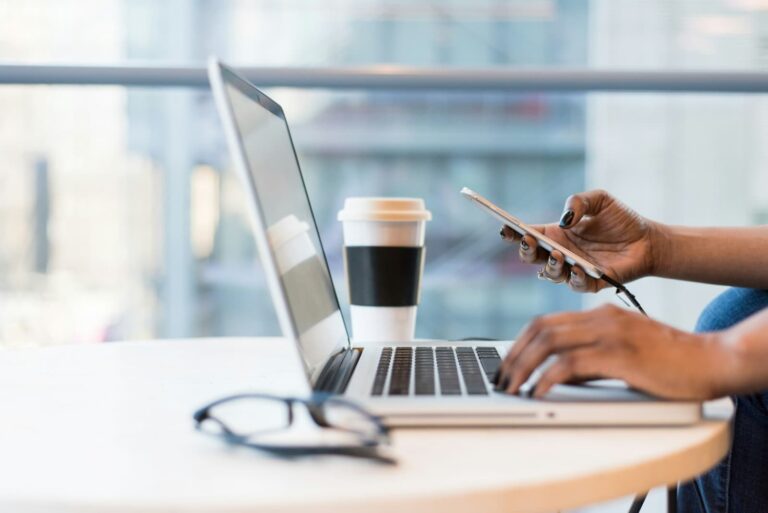 person sits at table typing on a laptop while scrolling on phone getting updates