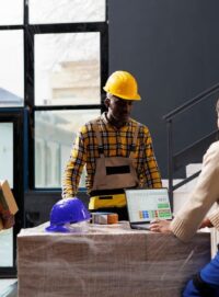 three employees working inside a warehouse with tall shelves of boxes and yellow hard hats working on a laptop