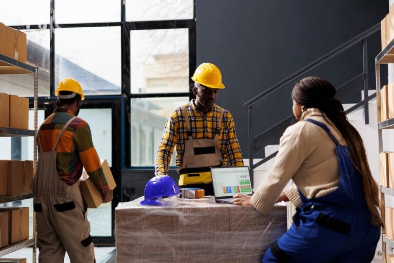 three employees working inside a warehouse with tall shelves of boxes and yellow hard hats working on a laptop