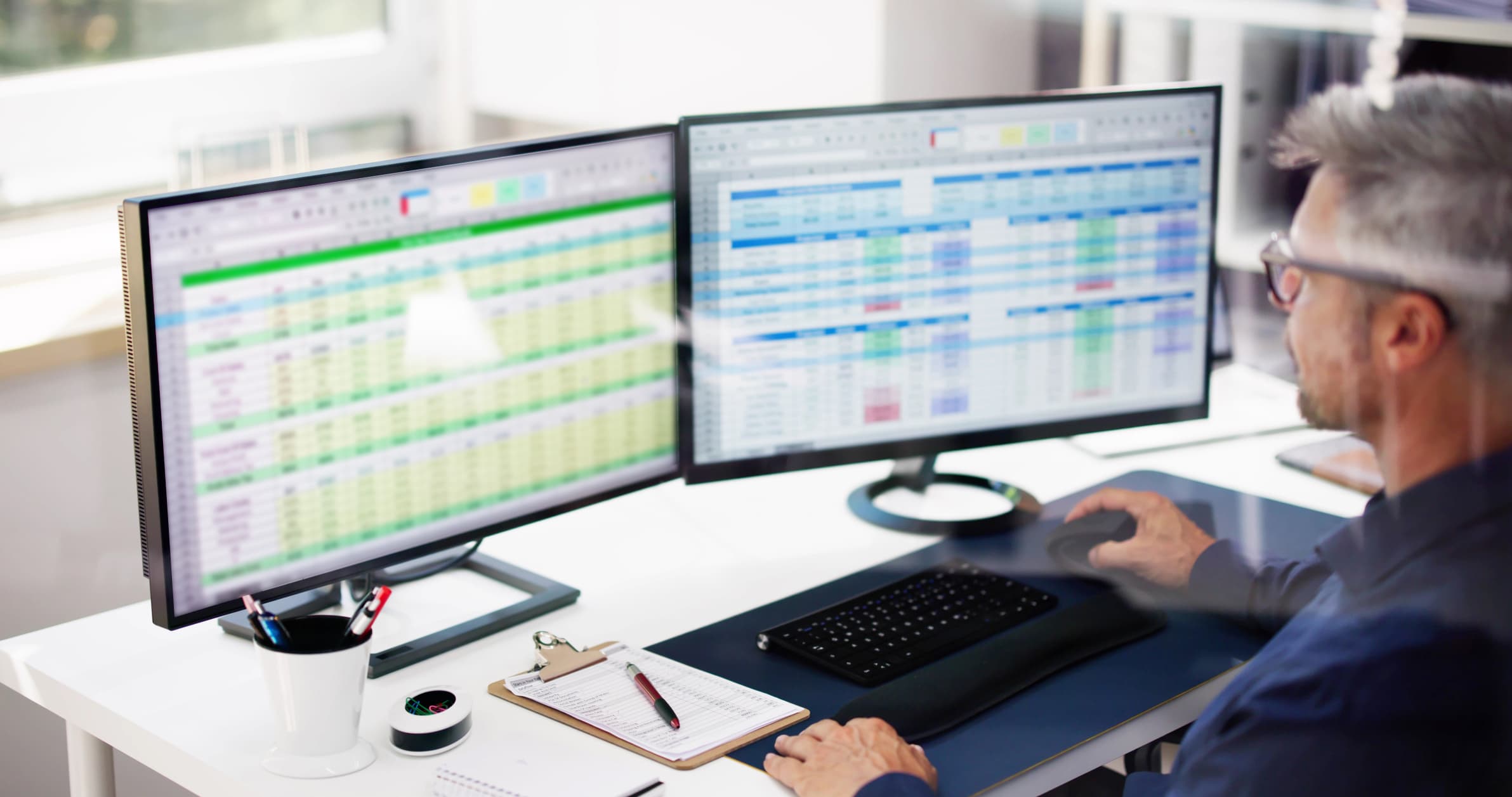 man sits at desk looking at two computer screens that show spreadsheets on each screen